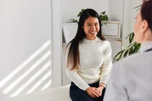 Woman with long dark hair in a white knit sweater sits in a bright office, smiling while talking to someone off-camera.