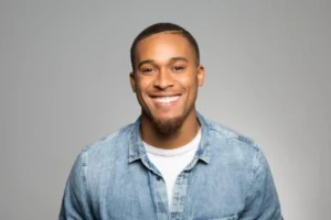 Smiling young man wearing a light denim shirt and white tee against a gray studio backdrop.
