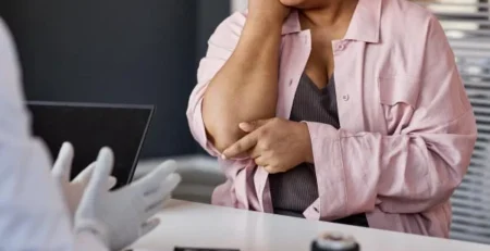 Woman in a pink shirt sits at a desk, touching her elbow in apparent pain during a medical appointment with a clinician in gloves.