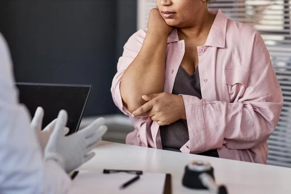 Woman in a pink shirt sits at a desk, touching her elbow in apparent pain during a medical appointment with a clinician in gloves.