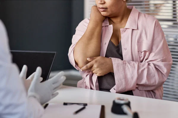 Woman in a pink shirt sits at a desk, holding her elbow in apparent pain during a medical consultation with a clinician wearing gloves across the desk closing the loop.