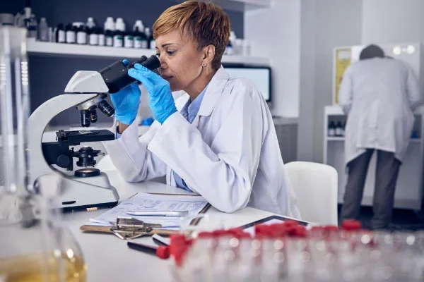 Female scientist in a white lab coat and blue gloves looks through a microscope at a lab bench, with test tubes and lab equipment nearby.