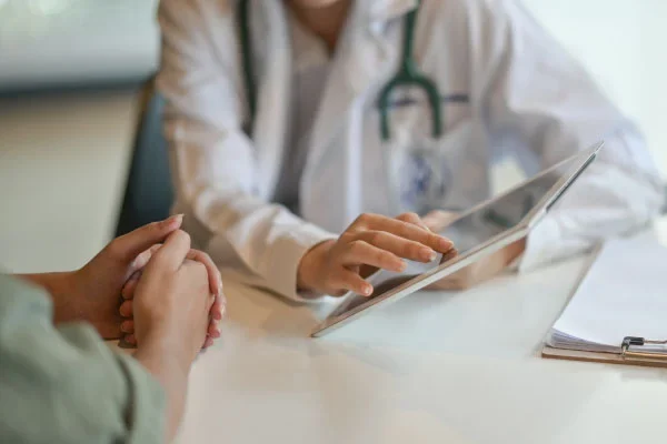 Doctor in white coat uses a tablet to discuss test results with a patient who has hands folded on the table.