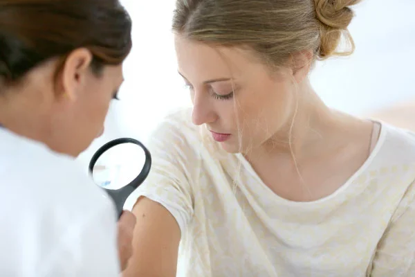 A clinician uses a handheld magnifying glass to inspect a woman's forearm during a medical examination.