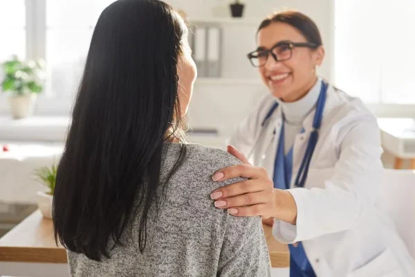 Doctor in a white coat with a stethoscope greets a patient, placing a reassuring hand on her shoulder as they smile at each other in a clinic.